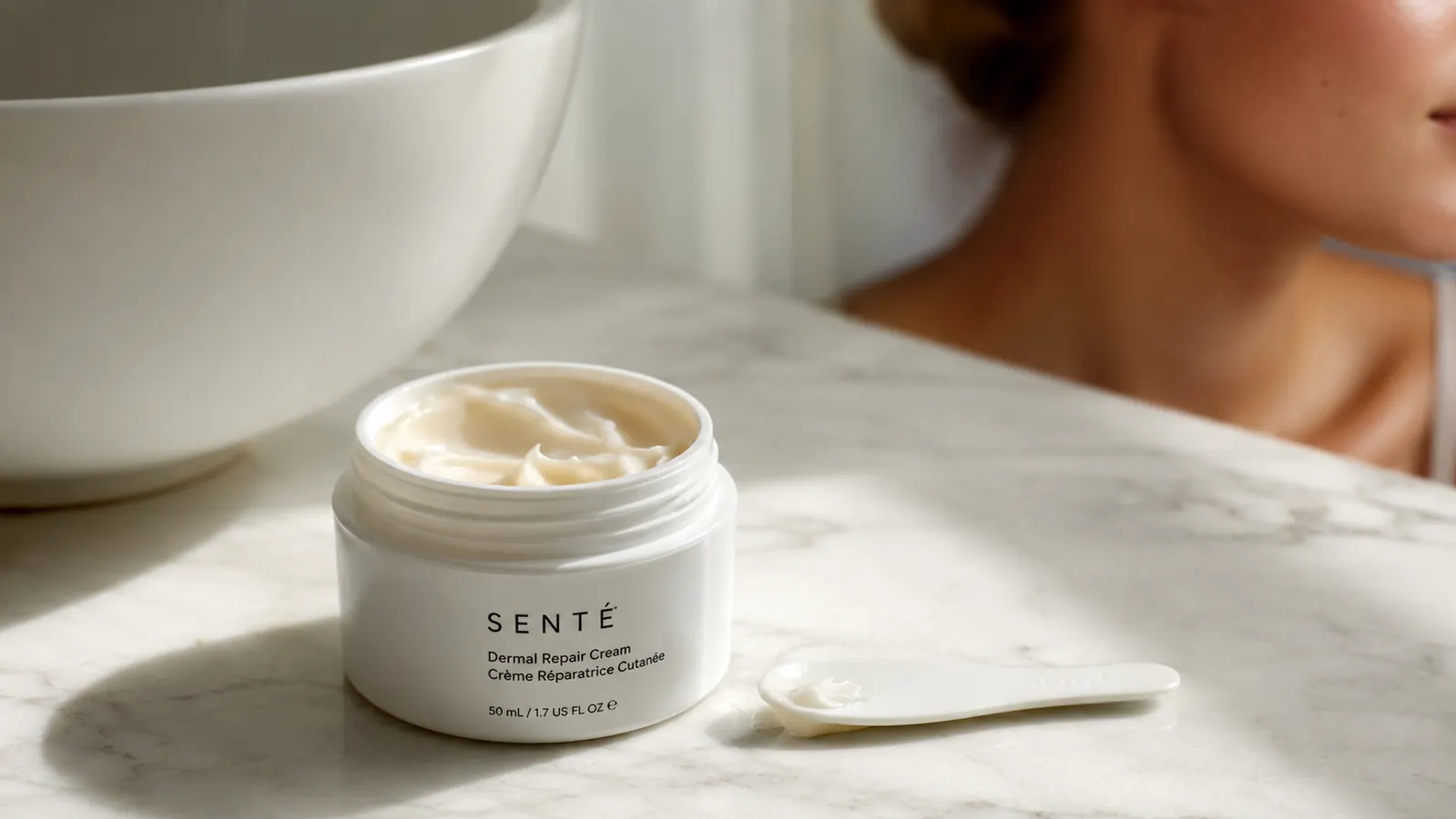 Close-up of a jar of white moisturizer being opened on a bathroom countertop with soft morning light coming through a frosted window