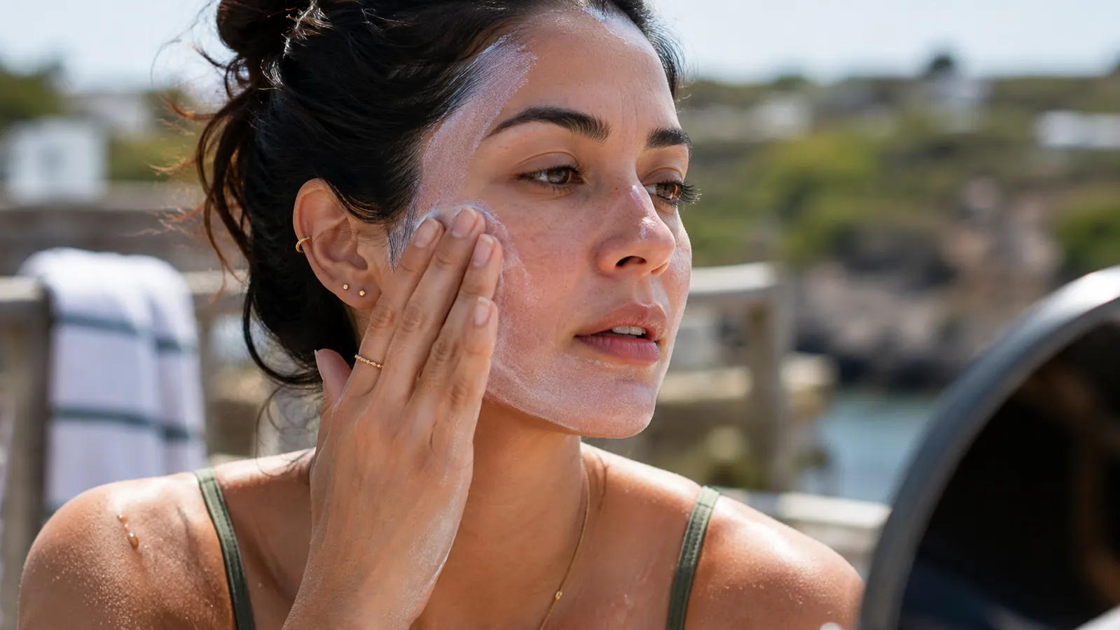 Person outdoors in sunlight rubbing sunscreen into their face, with a slightly white cast visible along the jawline