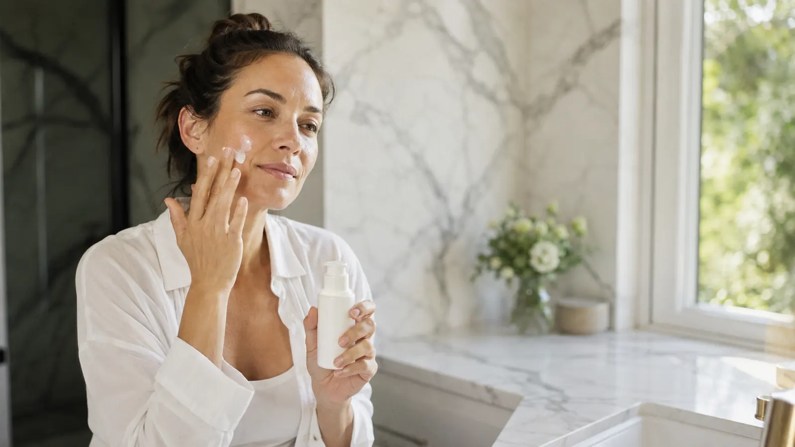 Person applying moisturizer to slightly damp skin at a bathroom sink, soft morning light