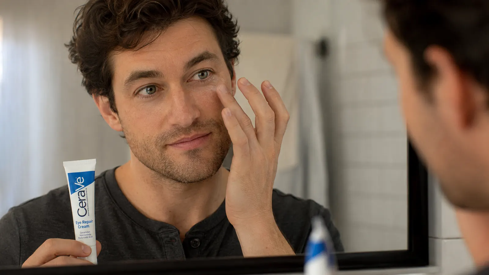 Man applying eye cream with his ring finger to the under-eye area in a bathroom mirror