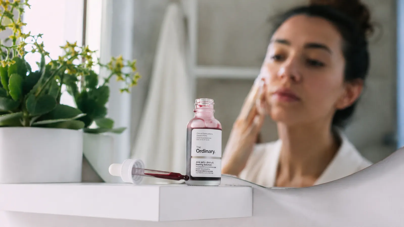 TruSkin Vitamin C serum bottle on a bathroom shelf beside a mirror showing a person examining their skin
