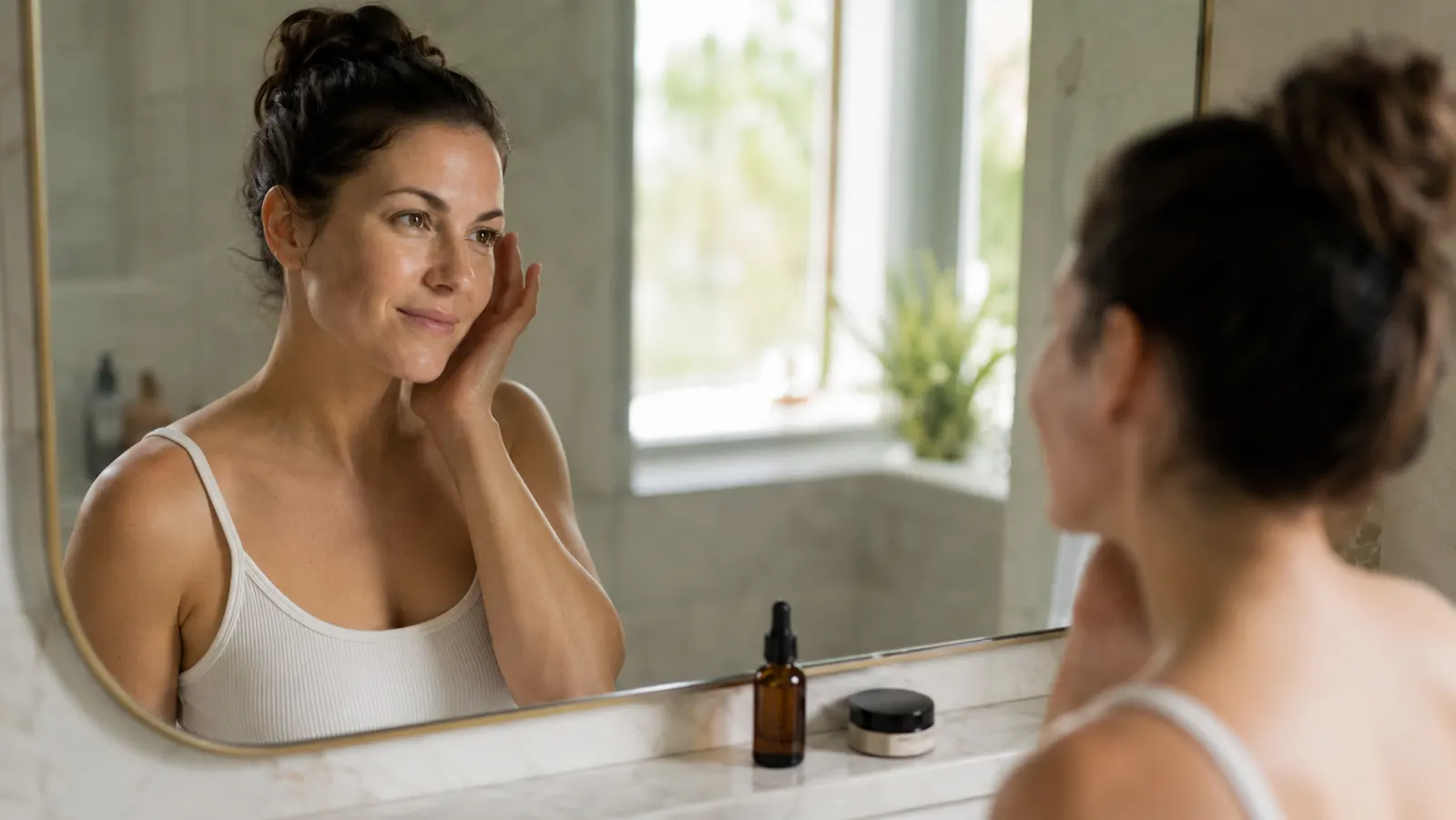 Woman looking at her reflection in a bathroom mirror, morning light, calm expression, clear skin
